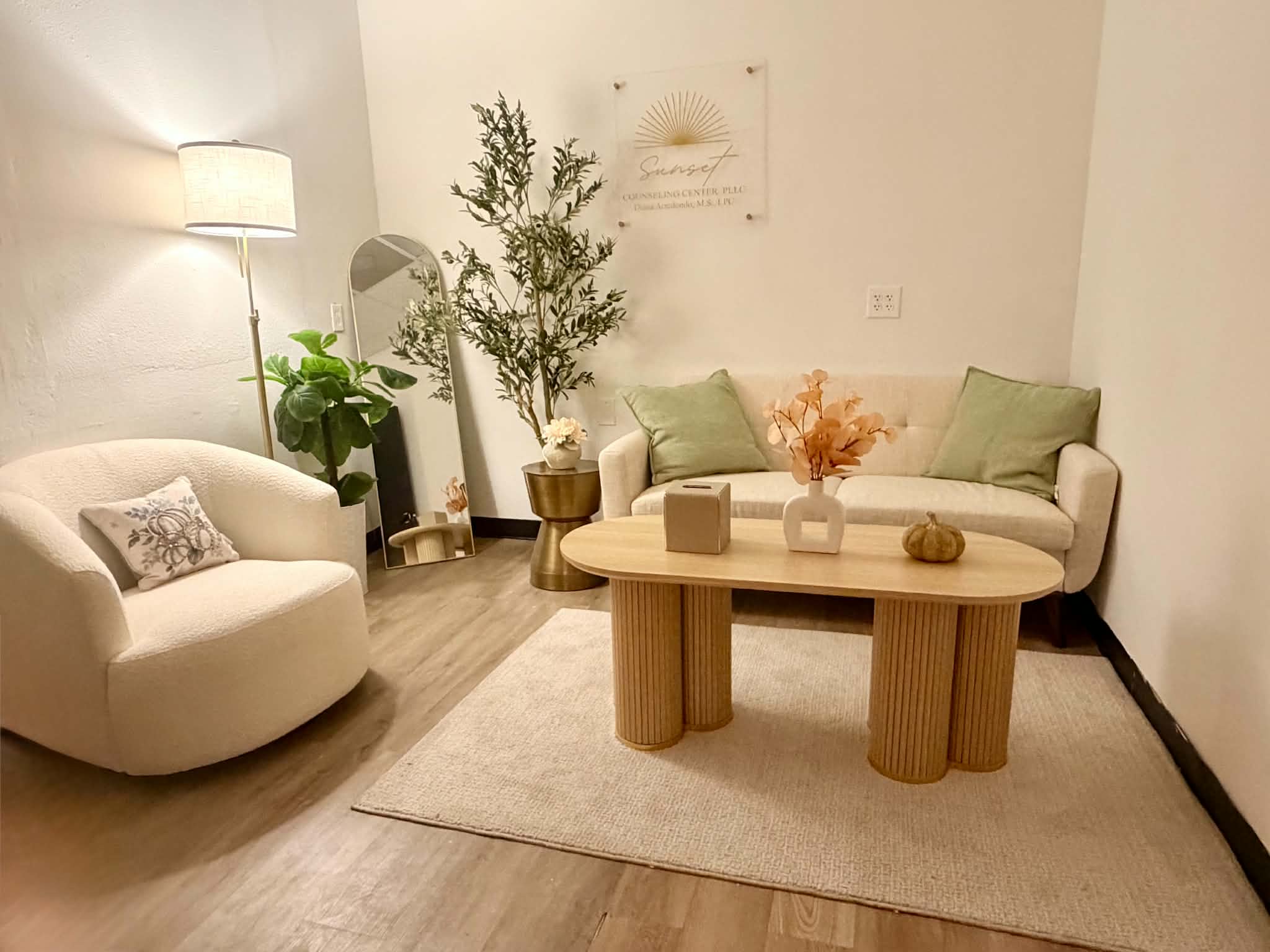 A calm counseling room at Sunset Counseling Center with a cream sofa, sage green pillows, a swivel armchair, an olive tree, and soft lamp light